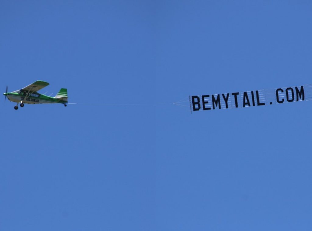 Aircraft with banner reading BEMYTAIL.COM towed behind
